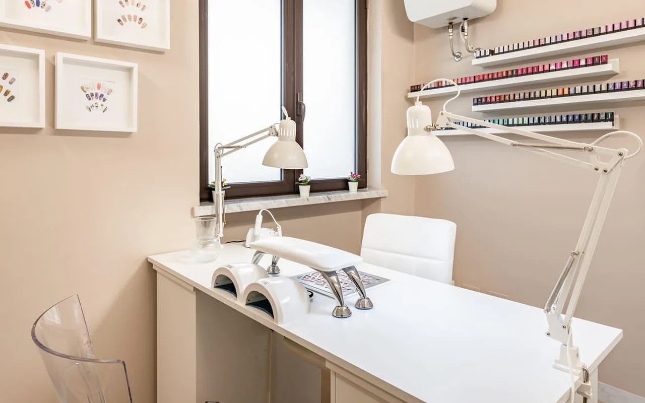 Eine eigene Nagelstation bei AG The Beauty Room's mit einem sauberen weißen Tisch, zwei UV-Lampen, verstellbaren Arbeitsleuchten und Regalen voller bunter Nagellacke. Gerahmte Nailart-Designs hängen an den warmtonigen Wänden.
