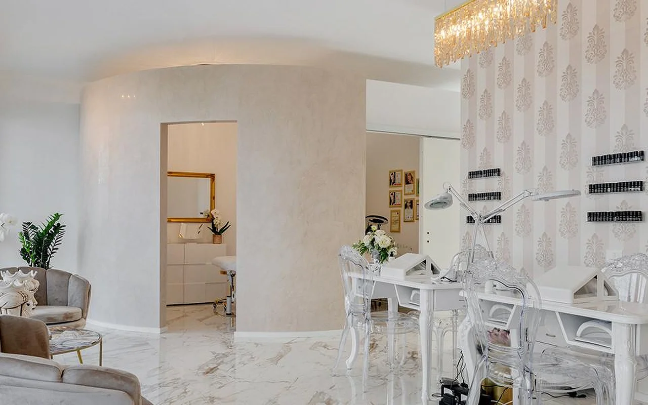 Das elegante Interieur des MC Beauty Center mit Boden in Marmoroptik, weißen geschwungenen Wänden und einem goldenen Kristall-Kronleuchter. Rechts steht eine Nagelstation mit Ghost-Chairs, durch eine goldgerahmte Tür gelangt man in einen Behandlungsraum dahinter.