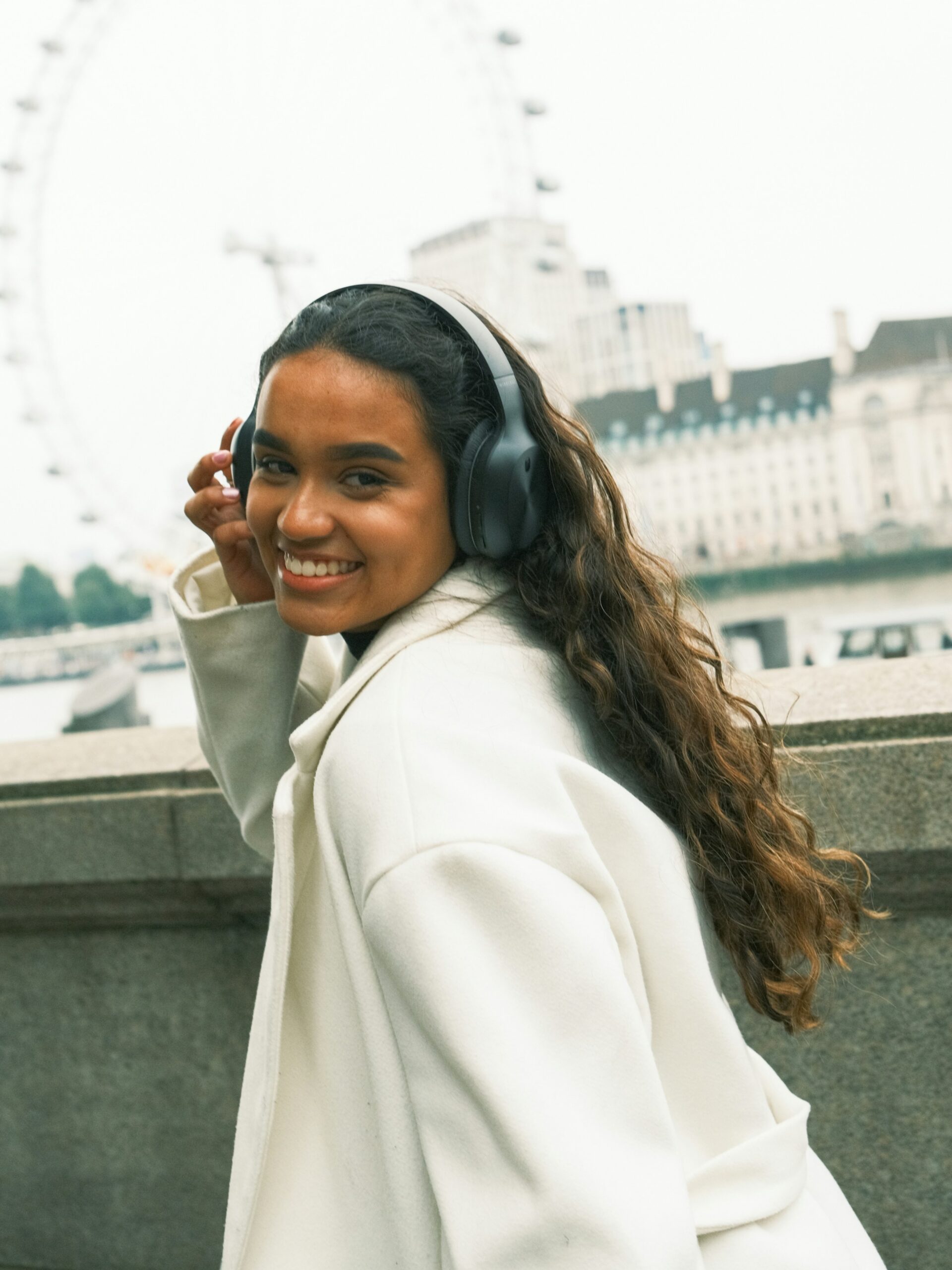 Eine lächelnde Frau mit Microblading an den Brauen, schwarzen Over-Ear-Kopfhörern und einem weißen Mantel auf einer Brücke