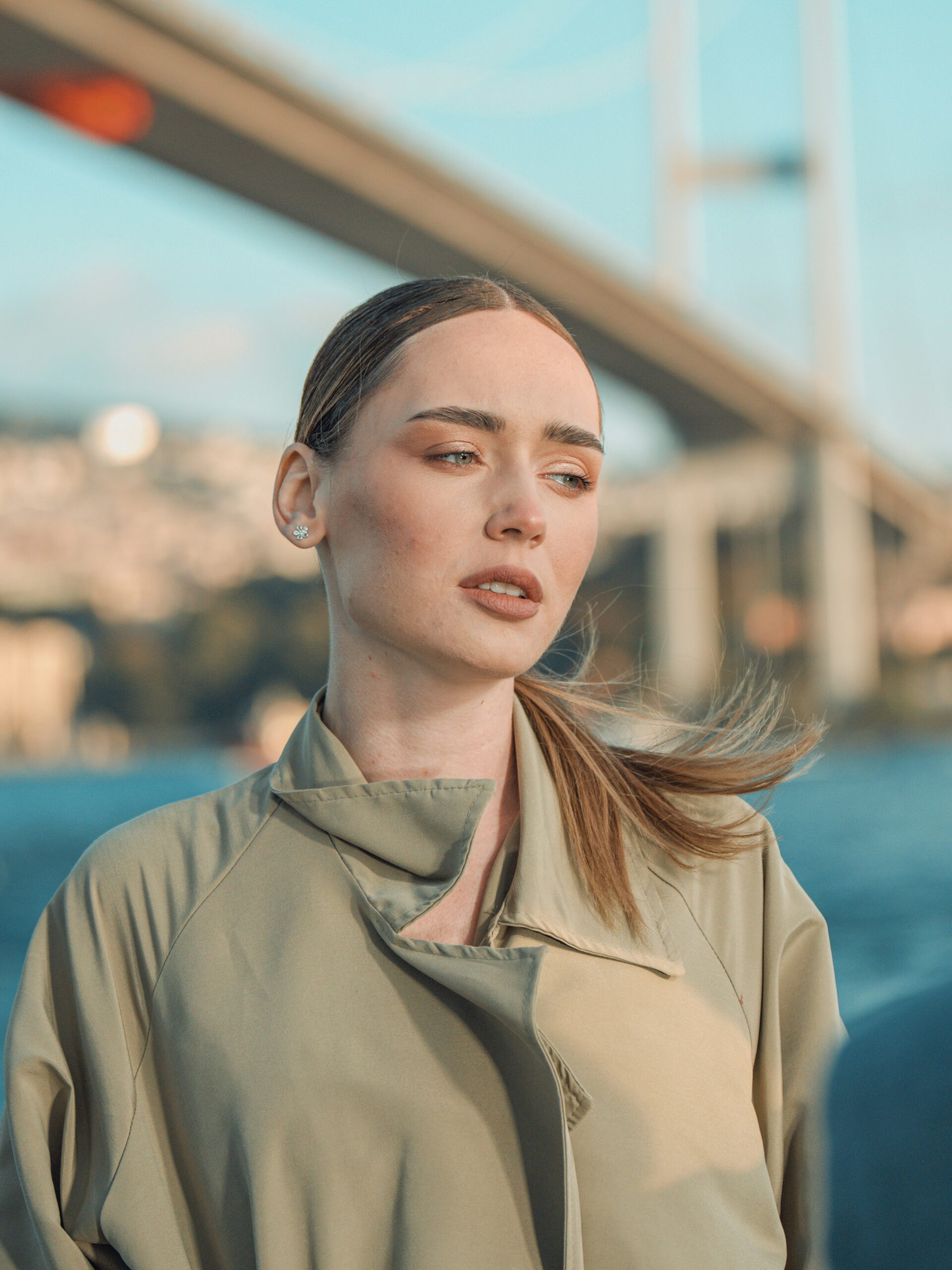 Eine Frau mit Microblading an den Brauen und hellem, zurückgebundenem Haar, im Trenchcoat vor einer Brücke in Istanbul