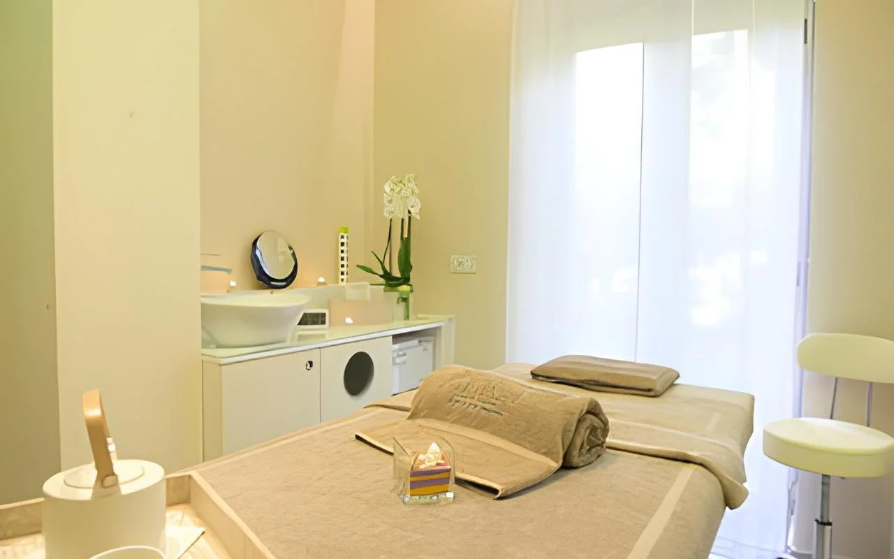 Ein ruhiger Behandlungsraum bei Alice Beauty Clinique mit einer Liege in weichem Taupe-Leinen, weißen Orchideen auf der Ablage, Kerzen am Waschbecken und transparenten Vorhängen, die das Tageslicht durch ein großes Fenster filtern.