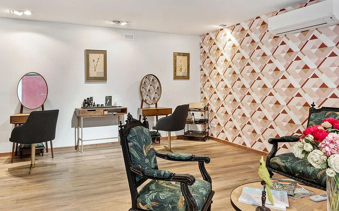Ein eleganter Salonraum mit einer geometrischen rosa-terrakottafarbenen Akzentwand, einem Paar kunstvoll geschnitzter Stühle mit grünem Botanik-Bezug, hölzernen Styling-Tischen mit runden Spiegeln und einer Vase mit rosa und roten Blumen im Vordergrund.