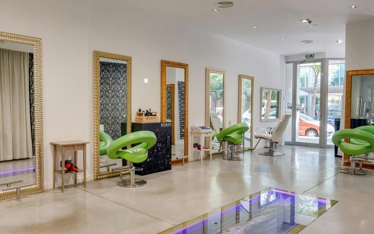 Ein heller, geräumiger Friseursalon mit poliertem Betonboden, hohen goldgerahmten Spiegeln an einer weißen Wand, limettengrünen geformten Styling-Stühlen und einem Glasboden-Bereich mit lila Licht, mit Blick auf die Straße durch verglaste Türen im Hintergrund.