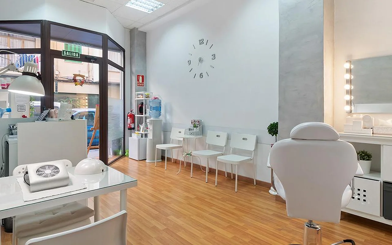 Ein heller, weißer Salon mit hellem Holzboden, einer Reihe weißer Wartestühle, einer minimalistischen Wanduhr, einem beleuchteten Schminkspiegel, einem weißen Behandlungsliegestuhl und einer Maniküre-Station am Fenster.