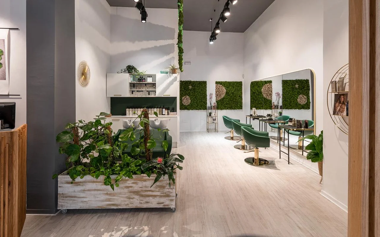 Ein pflanzengefüllter Friseursalon mit hellem Holzboden, einem gealterten weißen Übertopf voller Grün, grüner Moos-Wandkunst, smaragdgrünen Samt-Styling-Stühlen mit Golddetails und einer Waschstation dahinter.