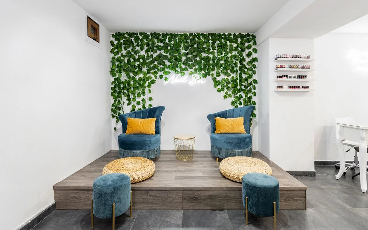 Eine Nagelstudio-Ecke mit einer herabhängenden grünen Blätterwand und einem kleinen Neonschild, zwei petrolfarbenen Samtsesseln mit senfgelben Kissen auf einer erhöhten Holzplattform, geflochtenen Hocker-Pouffes und Regalen voller bunter Nagellacke auf der rechten Seite.