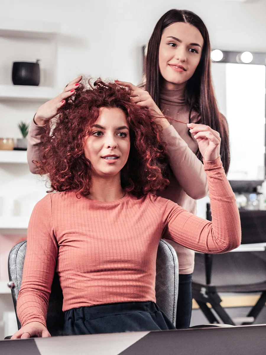 Eine Kundin mit rot gefärbtem, lockigem Haar sitzt beim Friseur, während ihre Stylistin die Behandlung mit ihr bespricht