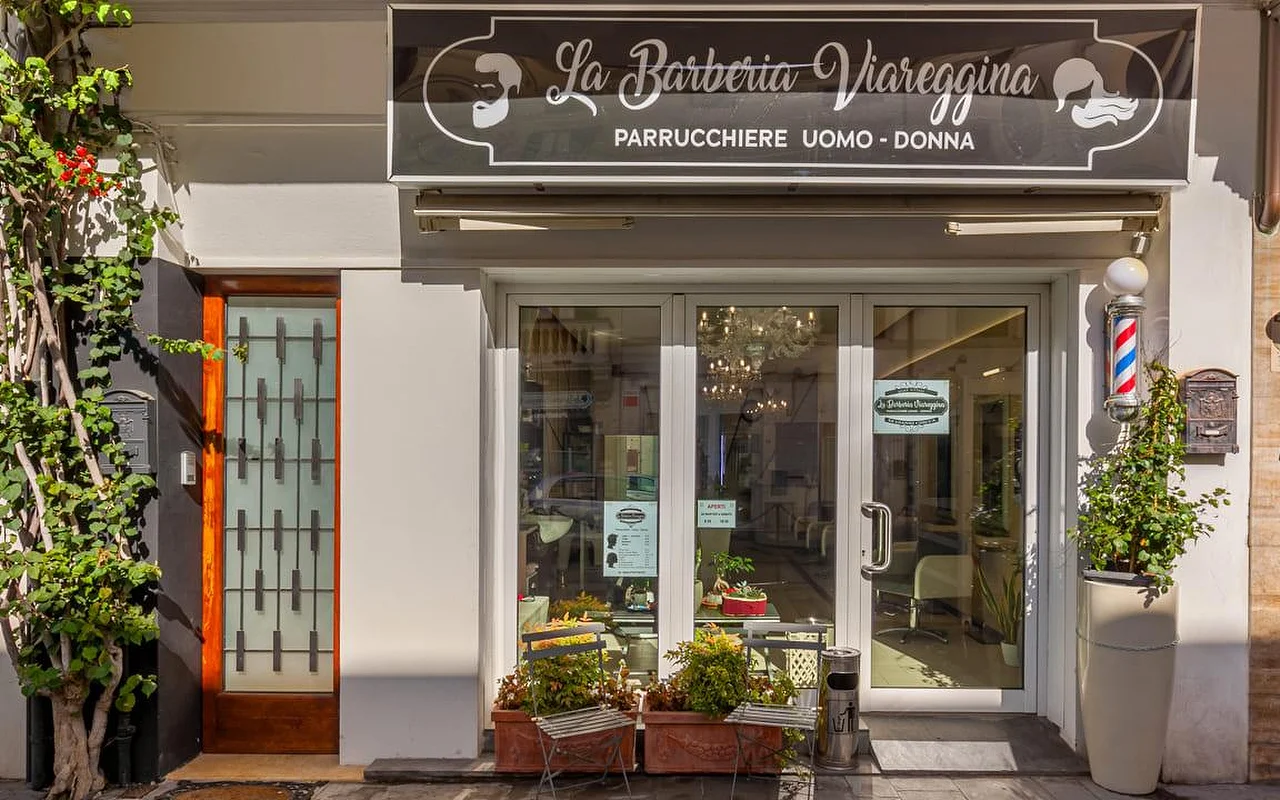 Der Eingang von La Barberia Viareggina an einem sonnigen Tag, mit einer klassischen rot-weiß-blauen Barber-Säule rechts neben den Glastüren. Topfpflanzen und rankendes Grün rahmen den Eingang, und durch die Fenster ist im Inneren ein Kronleuchter zu sehen.
