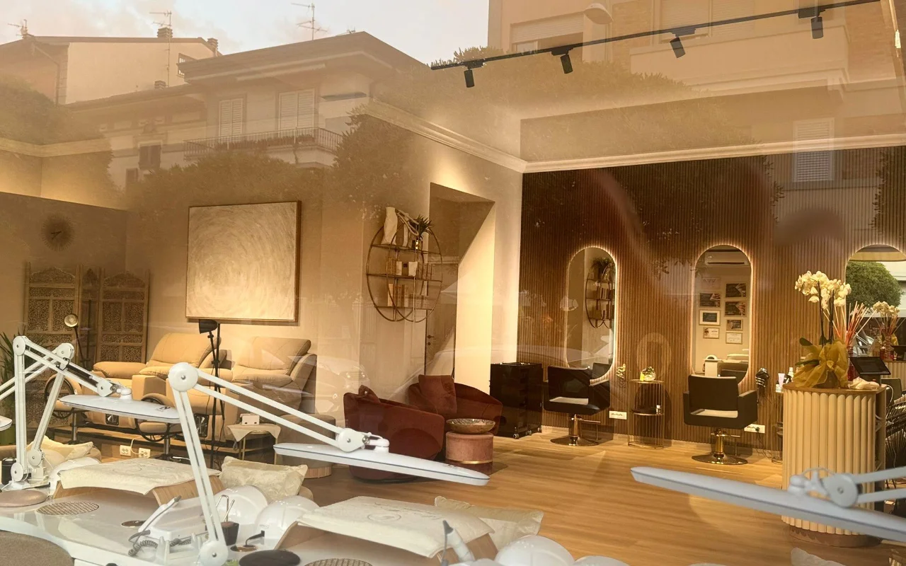 Das Interieur von Lilian Almanzas Salon, durch das Schaufenster zur goldenen Stunde gesehen. Nagelstationen mit verstellbaren Lampen säumen die linke Seite, rechts gibt es warme Holzvertäfelung, Bogenspiegel mit hinterleuchteten Rahmen und Samtsessel.