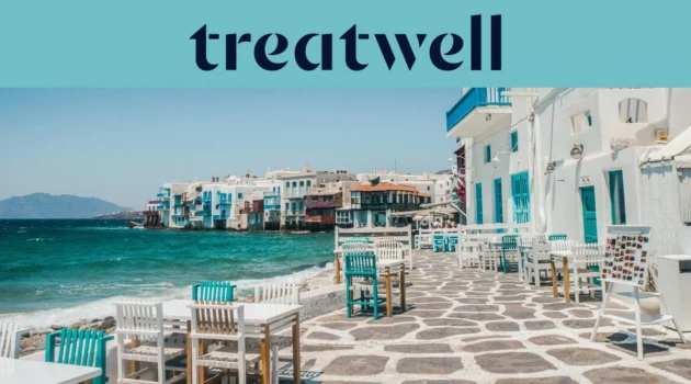 Die Uferpromenade von Klein-Venedig in Mykonos-Stadt: weiß getünchte Häuser mit bunten Holzbalkonen direkt am Wasser, dahinter die türkisfarbene Ägäis und im Vordergrund weiße und petrolfarbene Café-Tische auf einer steingepflasterten Terrasse.