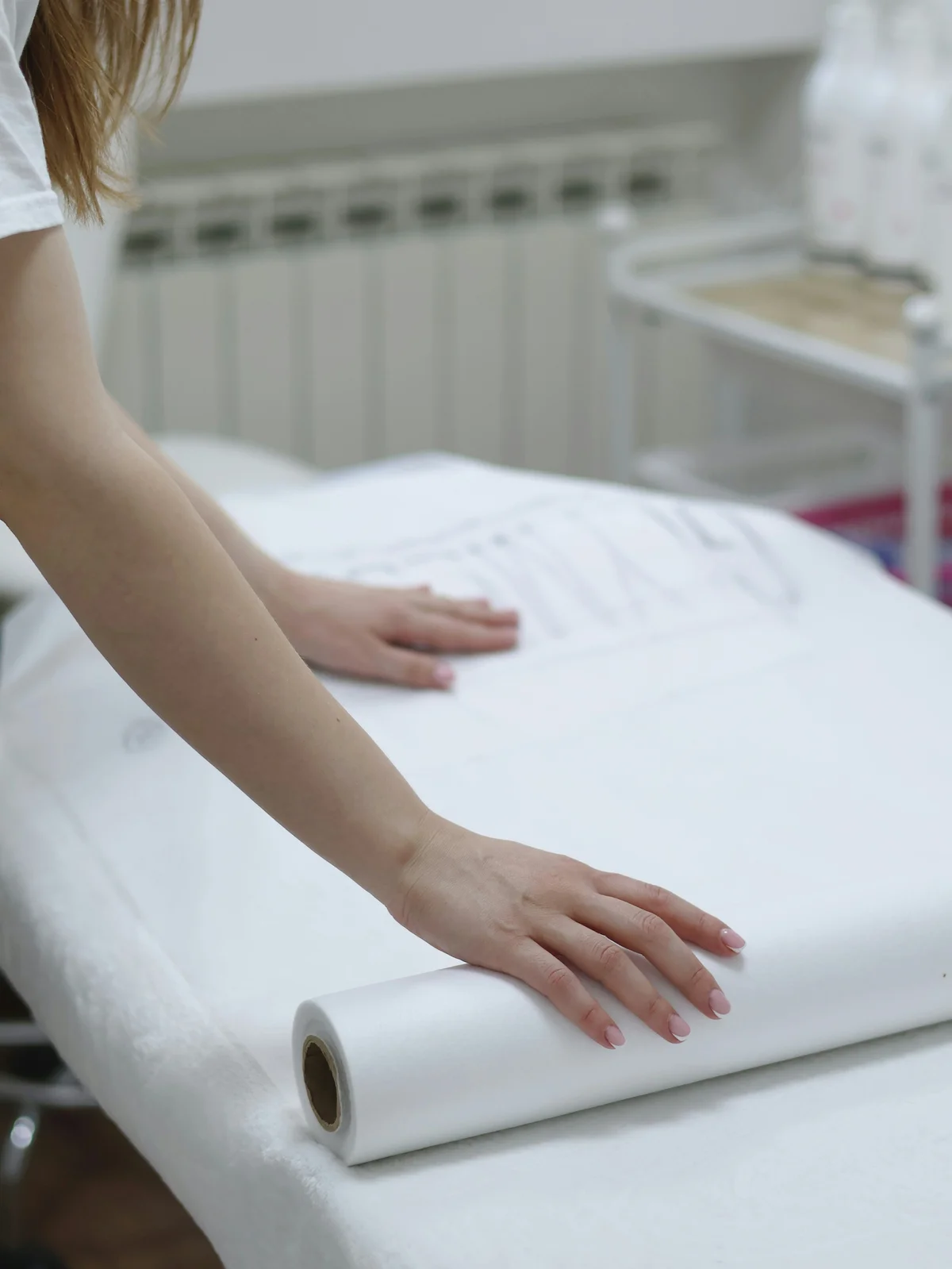 Nahaufnahme der Arme einer Kosmetikerin, die ein sauberes Blatt Papier auf einer Behandlungsliege ausrollt, um eine Behandlung vorzubereiten