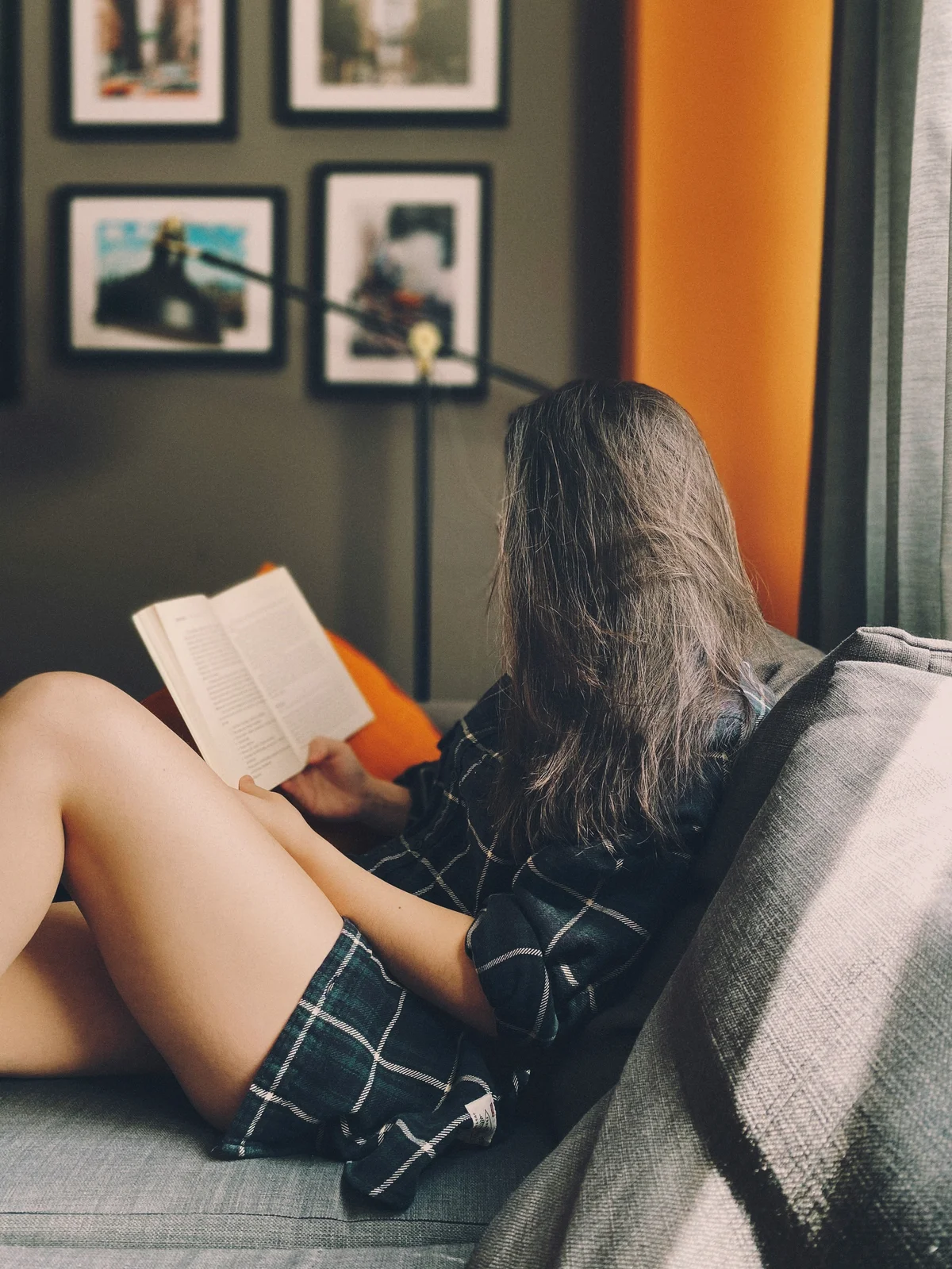 Seitenansicht einer Frau, die auf einem Sofa sitzt und ein Buch liest, während ihre langen Haare ihr Gesicht verdecken; sie trägt lockere Baumwollshorts und ein Hemd im dunkelgrünen Karomuster