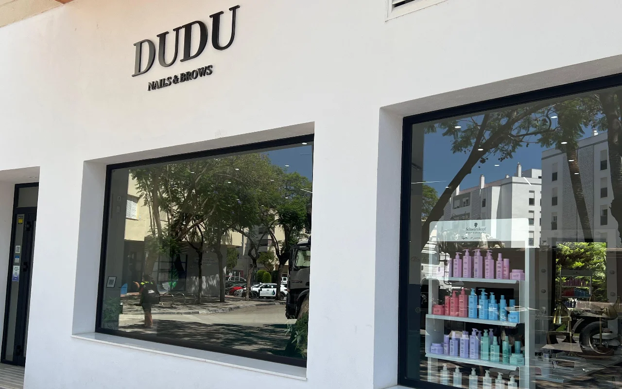 Die weiße Ladenfront von Dudu Nails and Brows in Estepona, der Name in dunklen Lettern über breiten Schaufenstern mit Reihen von Haarpflegeprodukten.