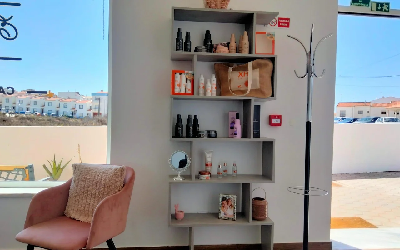 Eine Salonecke neben einem großen Fenster, mit einem gestuften grauen Regal voller Haarprodukte und einem gerahmten Foto, einem altrosa Samtsessel mit Kissen und Blick auf niedrige, weiß getünchte Häuser unter blauem Himmel.