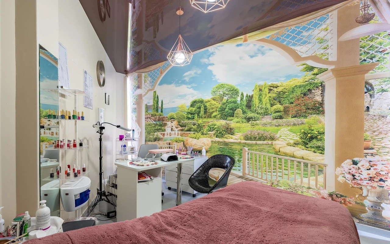 Ein Beauty-Behandlungsraum mit einer rosa drapierten Liege und einer weißen Nagelstation, vor einem großen Wandbild mit Garten und See, mit Glasregalen voller Produkte daneben.