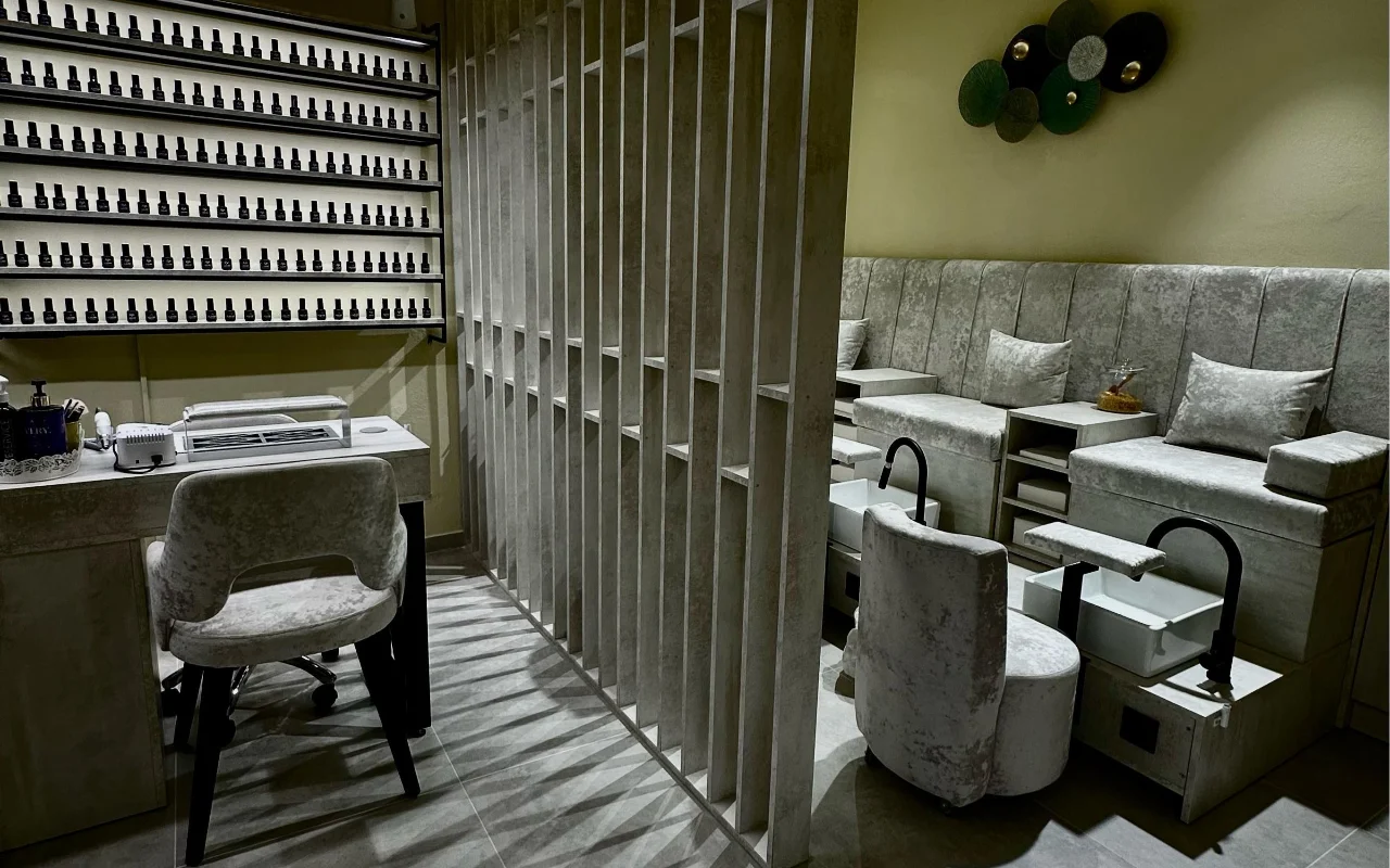 Ein Behandlungsraum bei Sisu Nail Club and Academy in Perissa auf Santorini, mit einer ganzen Wand voller Nagellacke, grauen Samtstühlen für Maniküre und Pediküre und einer durchbrochenen Betontrennwand.