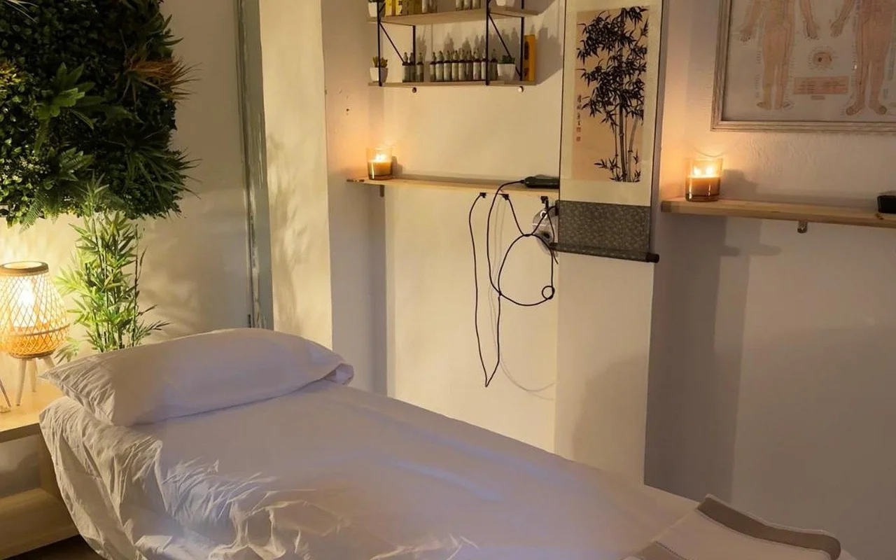 Ein schwach beleuchteter Behandlungsraum mit einer in weißem Leinen bezogenen Massageliege. Angezündete Kerzen stehen auf hölzernen Wandregalen neben einer Bambus-Tuschzeichnung und einer gerahmten Akupunktur-Tafel, mit einer Grünpflanze und einer leuchtenden Korblampe in der Ecke.